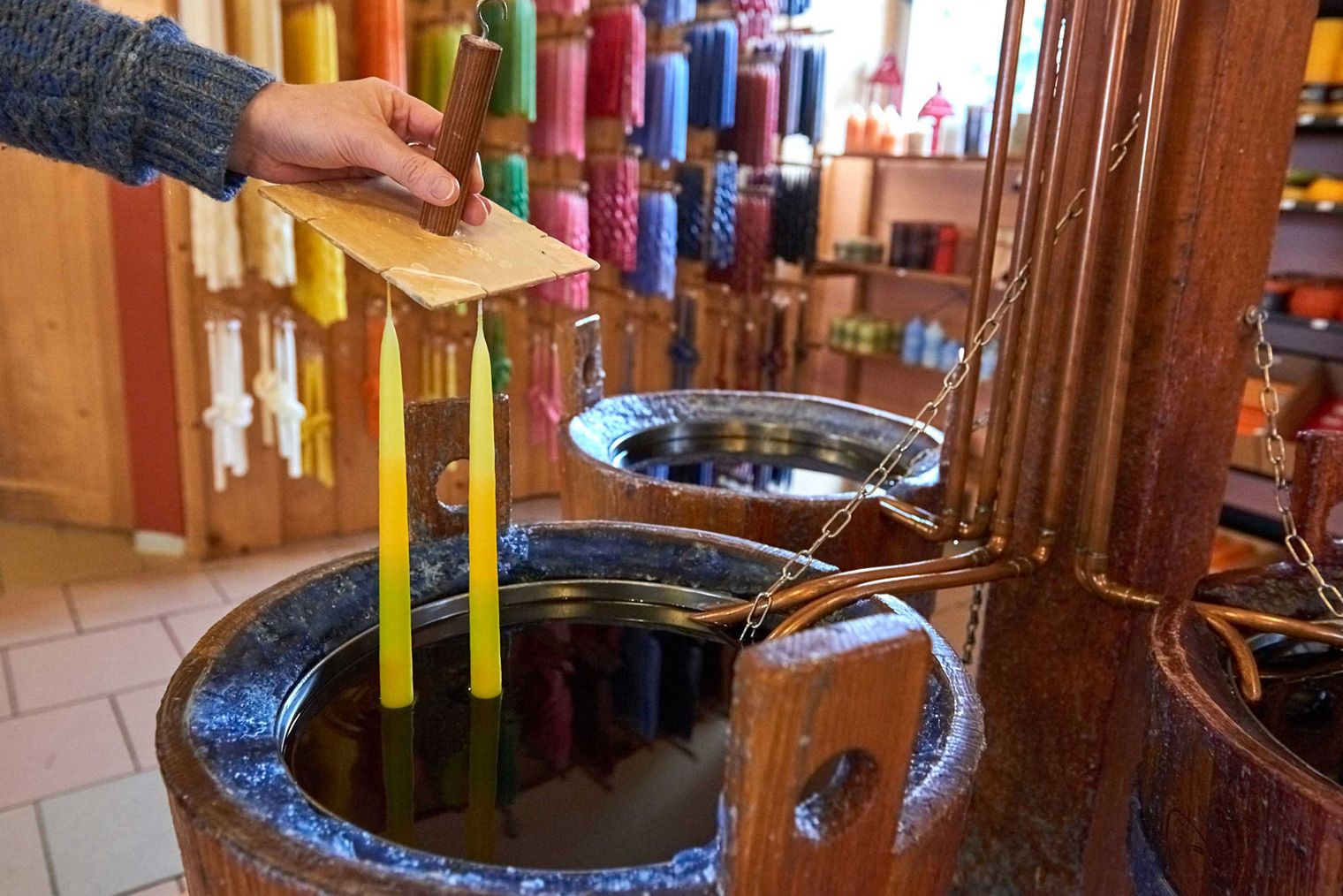 Hand taucht gelbe Kerzen in ein Gefäß mit Wachs, im Hintergrund bunte Kerzen.