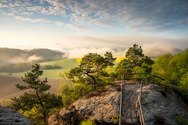 Felsformation mit grünen Baeumen, Weg entlang Gelaender auf Aussichtsplattform, blauer Himmel im Hintergrund. 