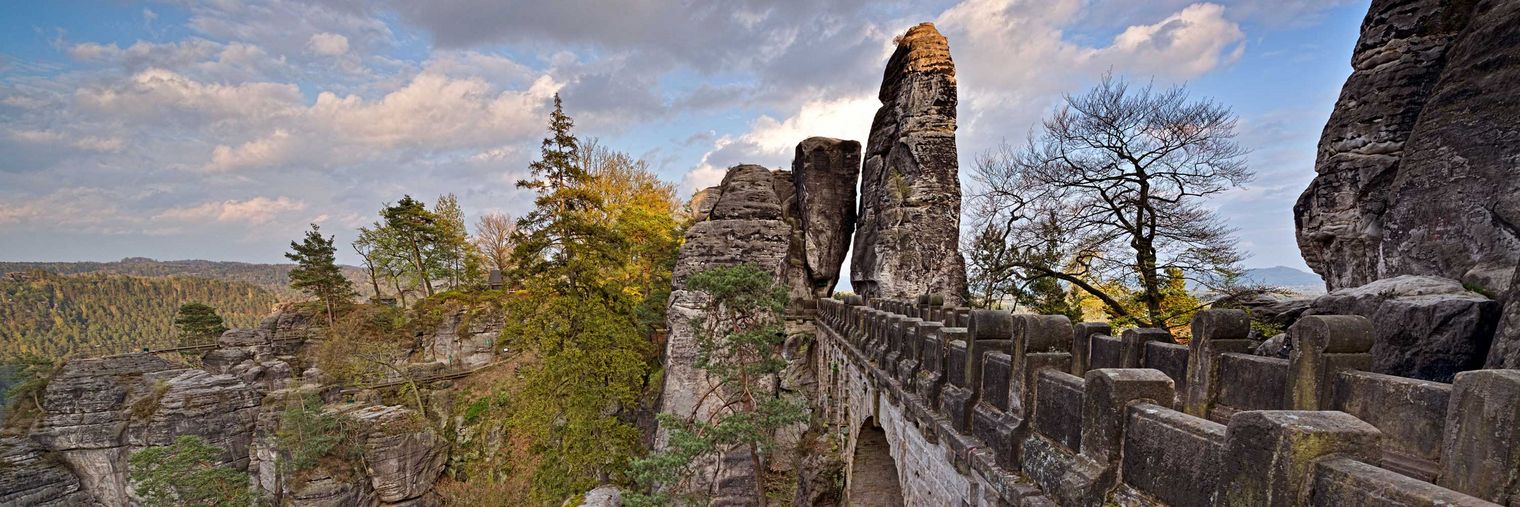 Brücke zwischen Felsen mit Bäumen und Wolken am Himmel, im Hintergrund das Felsentor Neurathen.