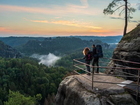 Person steht bei Felsenburg Bastei mit Sonnenaufgang, Nebel im Tal, spektakuläre Aussicht auf Felsen und Wälder.