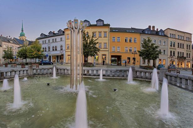 Marktplatz in Sebnitz mit Brunnen, bunten Gebäuden, und bewölktem Himmel.