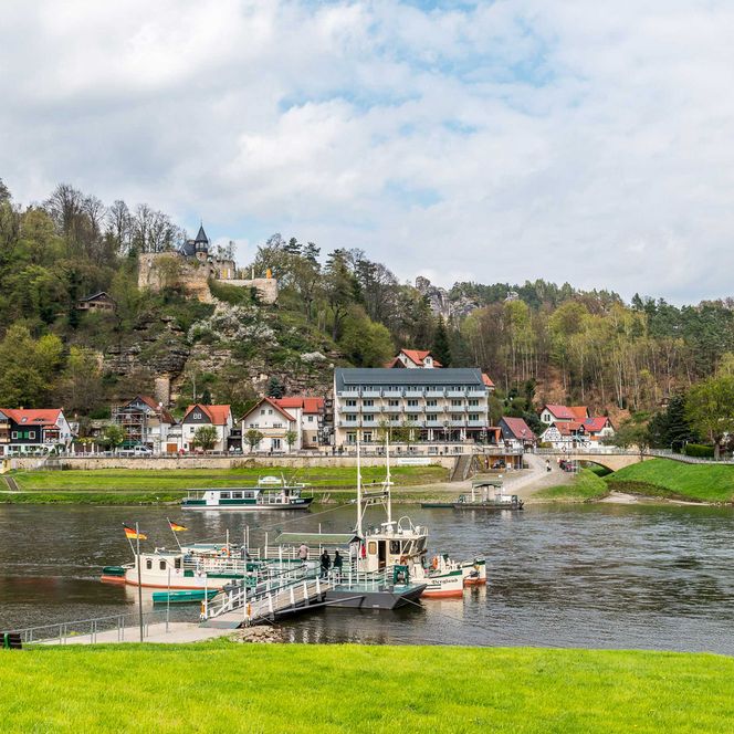 Blick auf einen ruhigen Fluss mit Booten und einer kleinen Stadt im Hintergrund, umgeben von Bäumen.