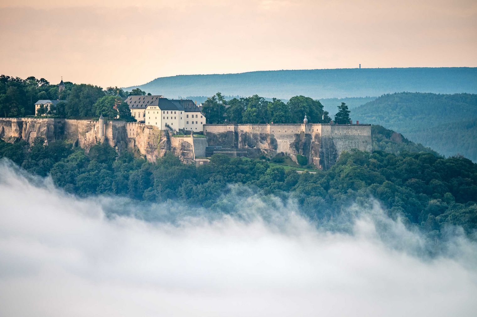 Burg auf einem Hügel, umgeben von Nebel und Bäumen, mit sanften Hügeln im Hintergrund.