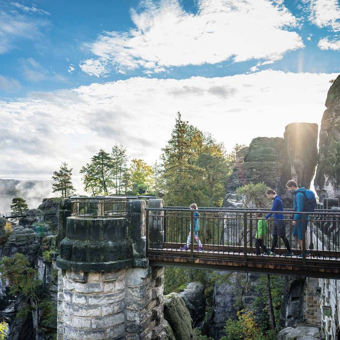 Blick auf eine Brücke mit Menschen, umgeben von Felsen und Bäumen, bei sonnigem Wetter.