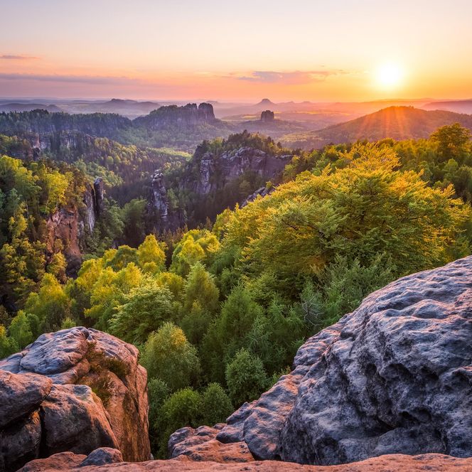 Sonnenaufgang über bewaldeten Bergen und Felsen, mit sanften Hügeln im Hintergrund.