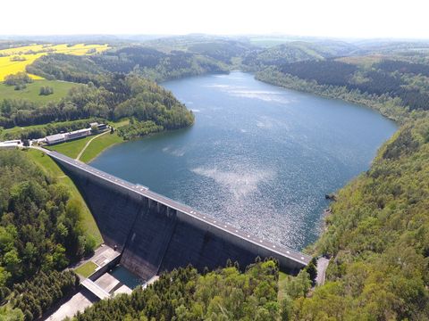 Talsperre von oben gesehen, umgeben von Wald, blauer Himmel mit Wolken, ruhige Wasseroberfläche.