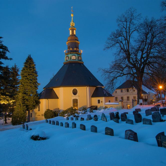 Kirche mit spitzem Turm, umgeben von schneebedecktem Friedhof und Bäumen, bei Dämmerung beleuchtet.