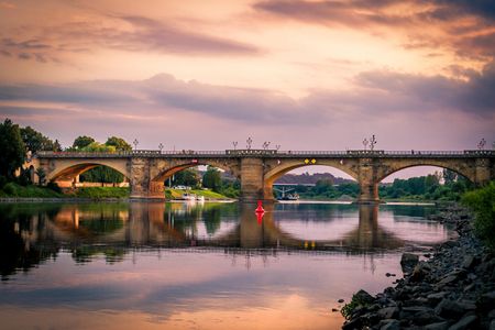 Brücke über einen ruhigen Fluss bei Sonnenuntergang, mit reflektierenden Farben im Wasser.