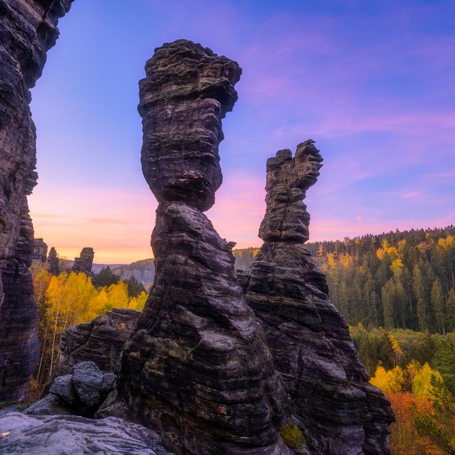 Felsformationen mit markanten, säulenartigen Strukturen vor einem bunten Sonnenuntergang und bewaldeter Landschaft.