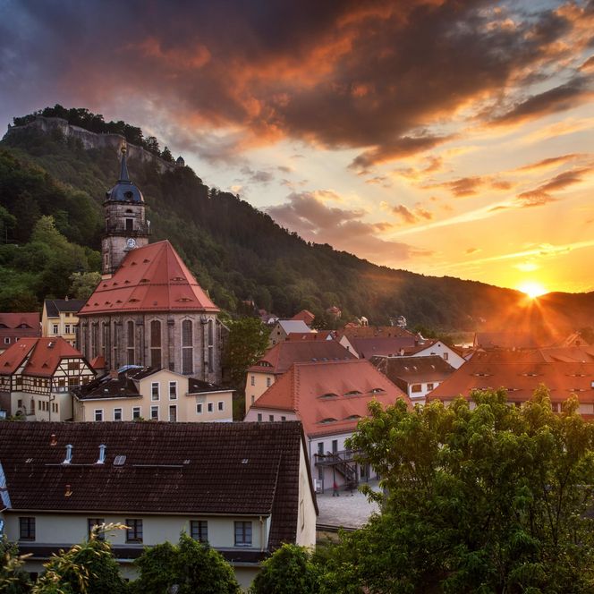 Sonnenuntergang über einer Stadt mit roten Dächern und einer Kirche, umgeben von Bergen und Bäumen.