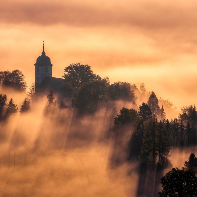 Blick vom Papststein auf die Kirche, umgeben von Nebel und orangefarbenem Licht des Sonnenaufgangs.