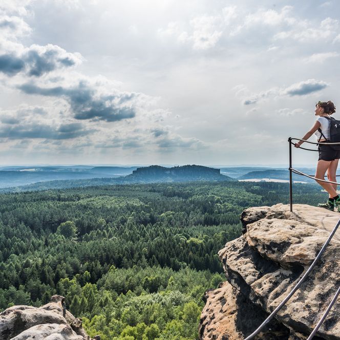 Wanderer mit Rucksack steht auf einem Felsen und blickt über eine bewaldete Landschaft unter einem leicht bewölkten Himmel.