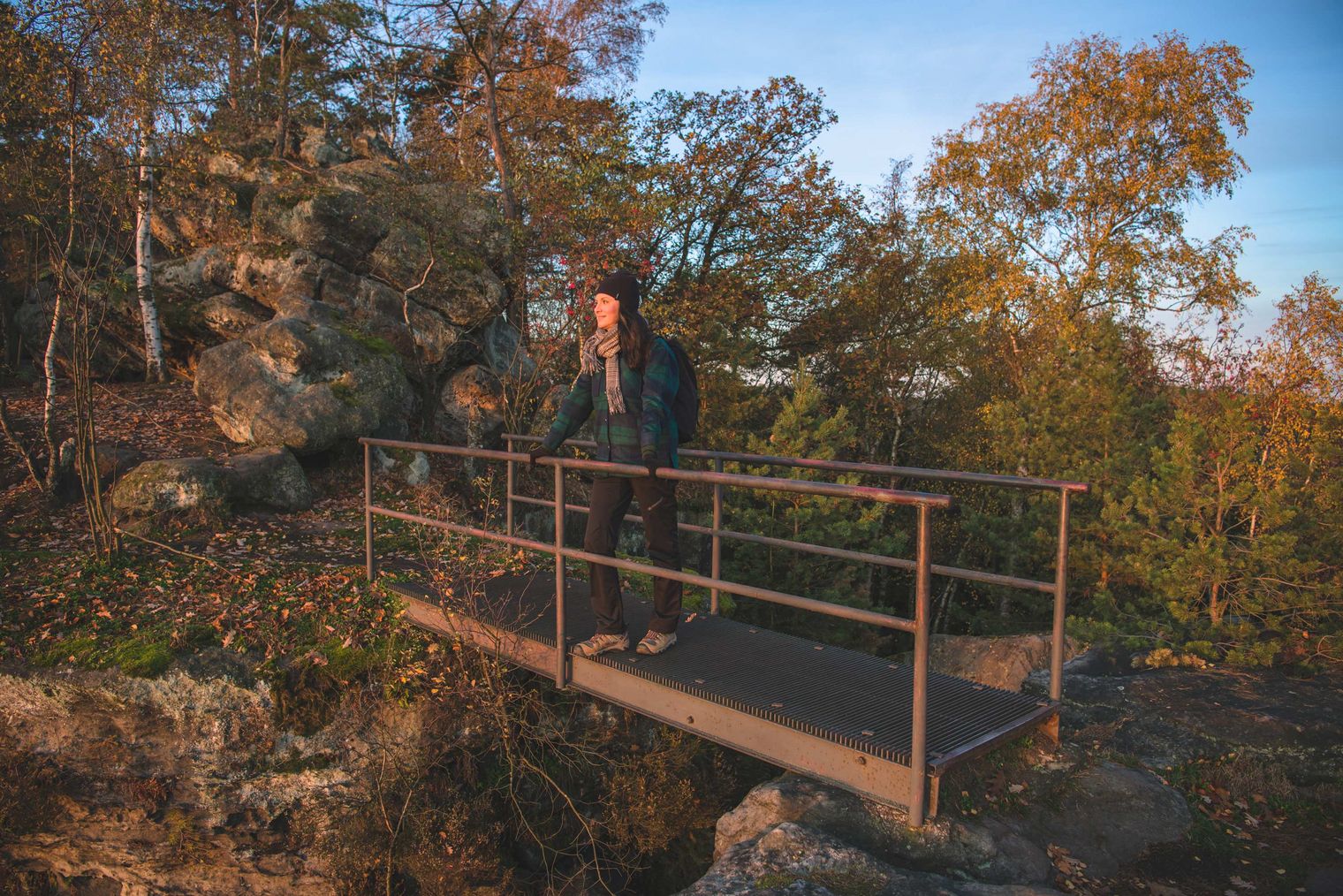 Person steht auf einer kleinen Brücke, über einer Felsspalte umgeben von Bäumen und Felsen, bei Sonnenuntergang.