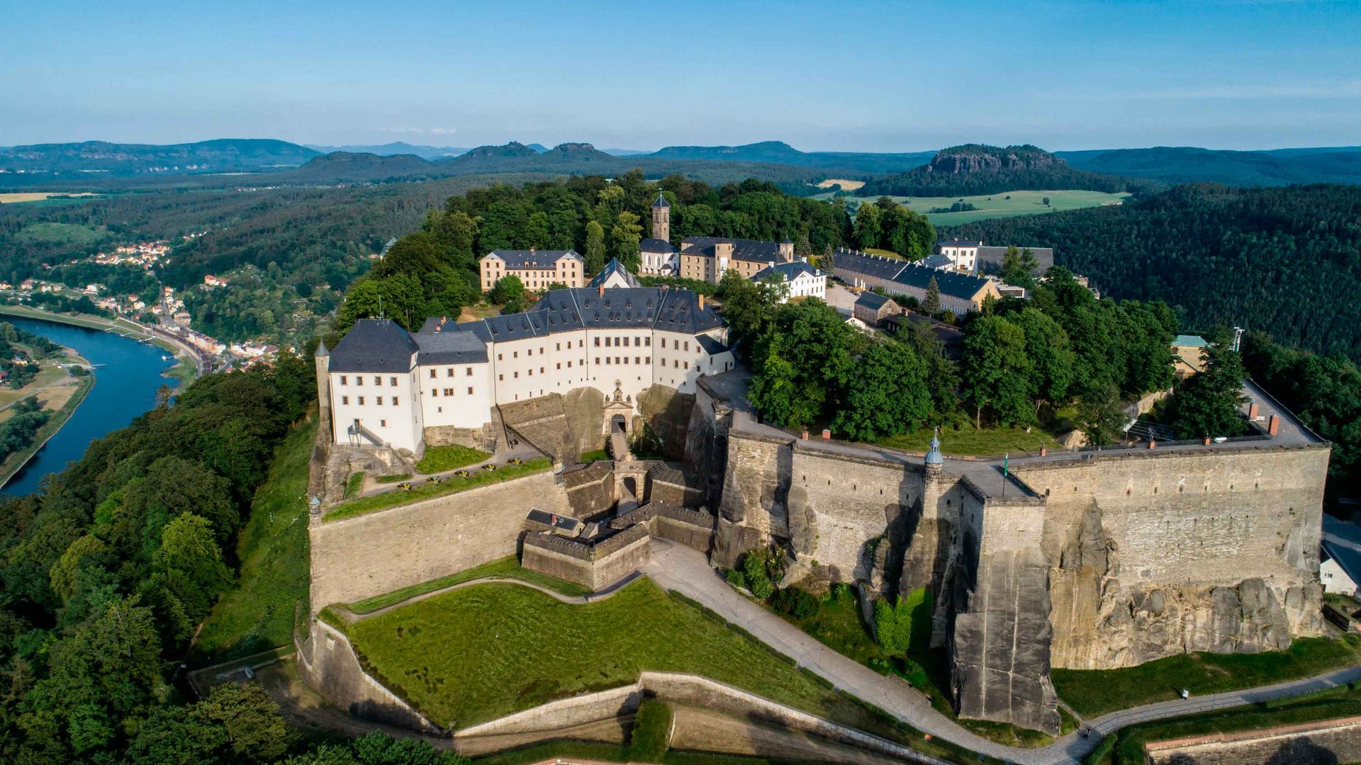 Festung Königstein: Luftaufnahme einer Burg auf einem Hügel, umgeben von grünen Wäldern und einem Fluss im Hintergrund.
