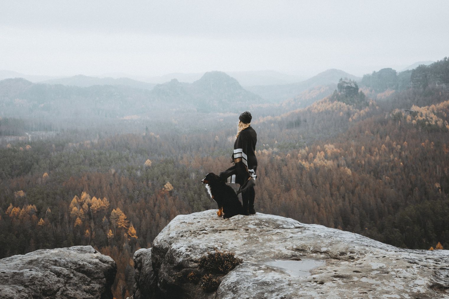 Frau steht mit einem Hund auf einen Felsen und schaut in die Ferne. Es ist nebelig.