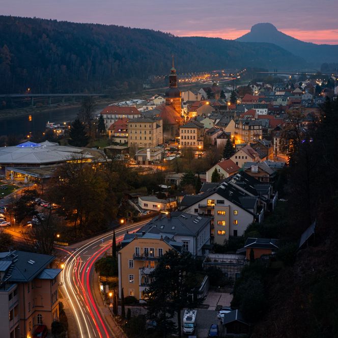 Blick auf eine Stadt bei Dämmerung mit beleuchteten Gebäuden und einem Berg im Hintergrund.