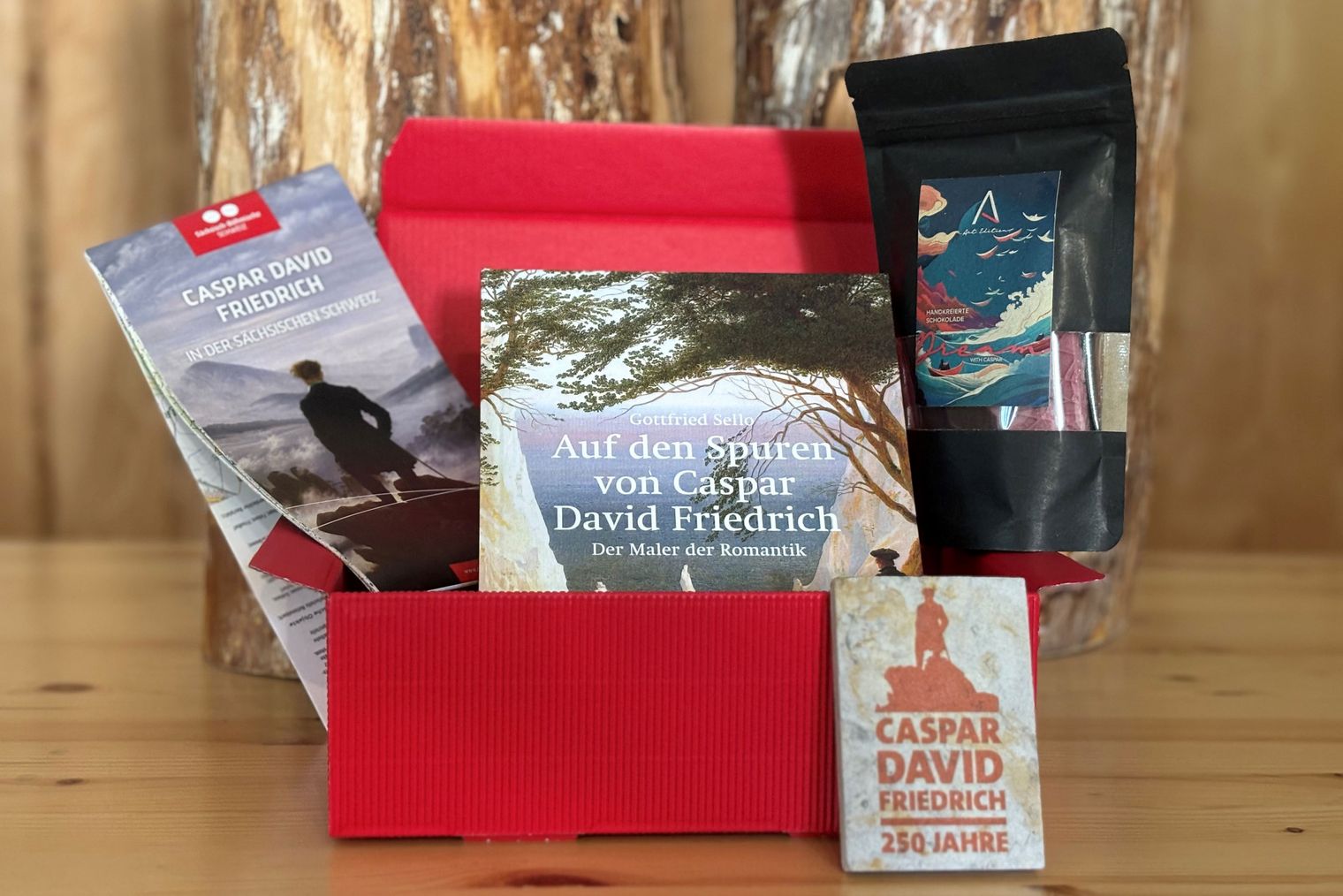 Rote Geschenkbox mit einer Karte, einem Buch, einer Tüte Schokolade und einem kleinen Magnet davor.