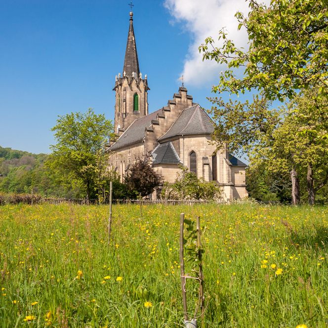 Kirche mit spitzem Turm, umgeben von grünem Gras und Bäumen unter blauem Himmel.