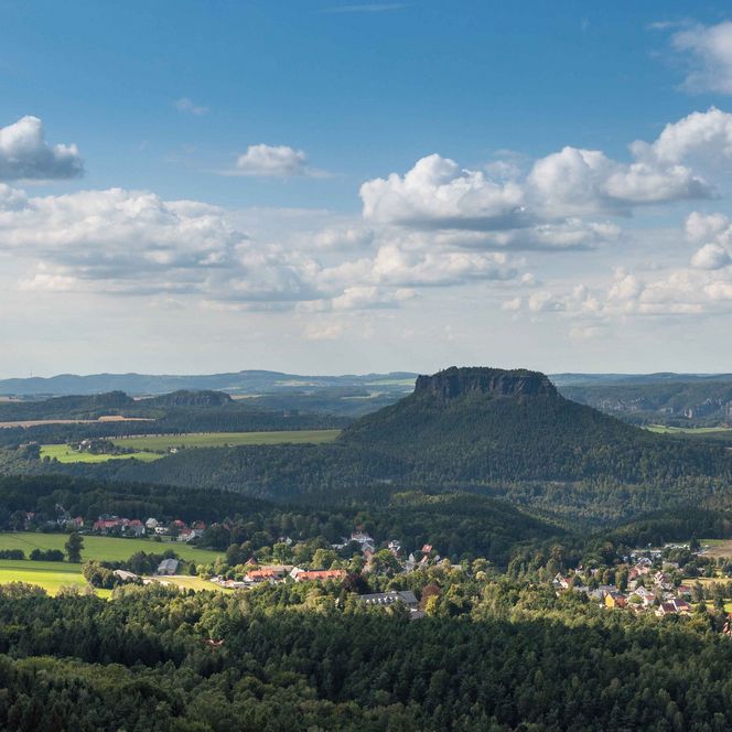 Blick auf eine grüne Landschaft mit Hügeln, Wolken und einem kleinen Dorf Gohrisch im Vordergrund.