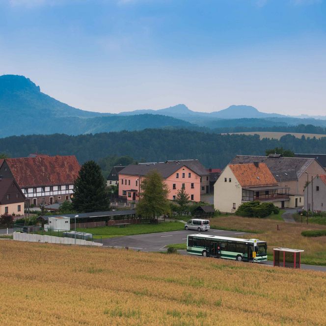 Landschaft mit einem Dorf, Feldern und Bergen im Hintergrund unter einem bewölkten Himmel.