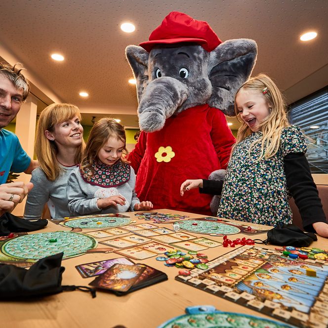 Familie spielt ein Brettspiel am Tisch, ein Elefantenmaskottchen steht daneben.