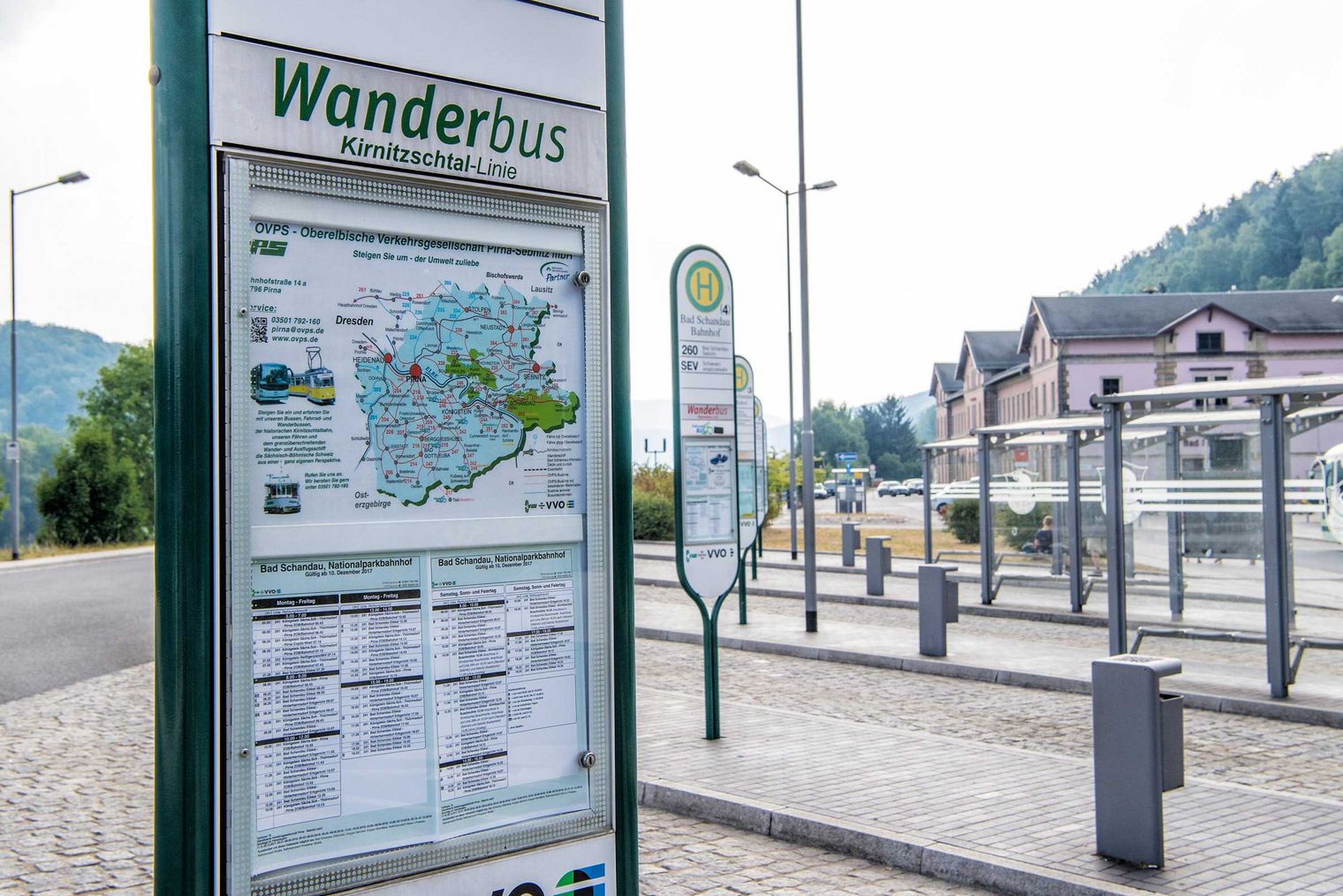 Wanderbus-Haltestelle mit einem Informationsschild und einer Karte der Umgebung. Berge im Hintergrund.
