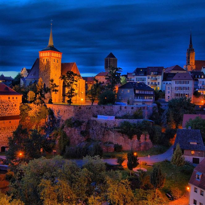 Blick auf eine beleuchtete Stadtmauer und Türme bei Dämmerung, umgeben von Bäumen und Häusern.