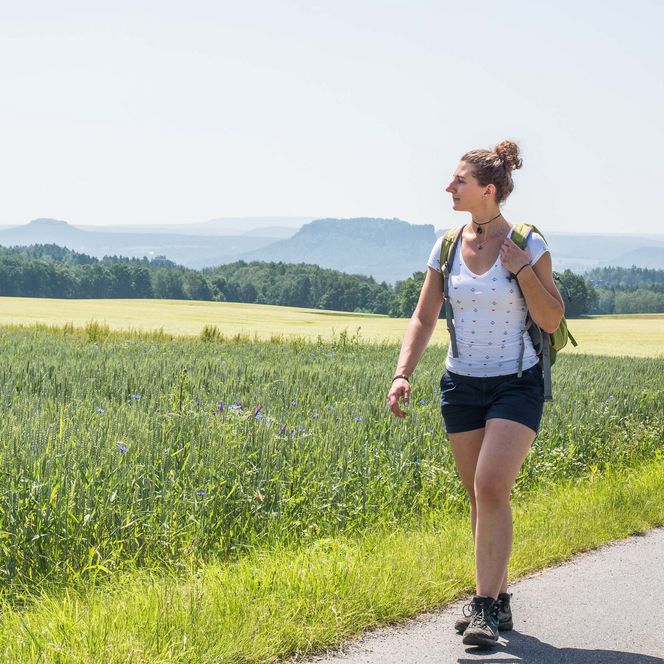 Junge Frau mit Rucksack wandert auf einem Weg durch eine grüne Landschaft mit Felsenlandschaft im Hintergrund.