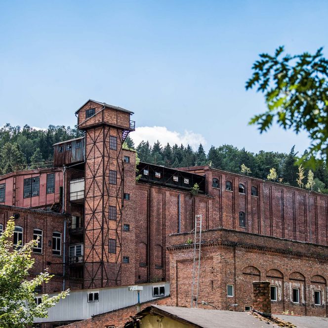 Rote Backsteinfabrik mit Türmen, umgeben von Bäumen und Hügeln unter blauem Himmel.