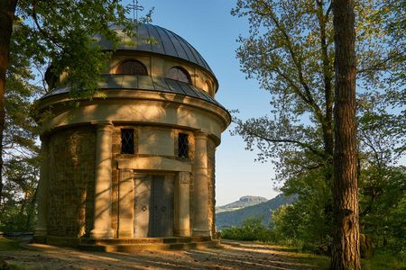 Malerwegkapelle Struppen Thürmsdorf: Runde Struktur mit einem kuppelförmigen Dach, umgeben von Bäumen und Bergen im Hintergrund.