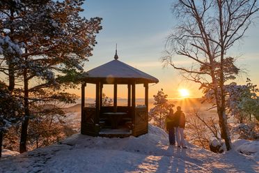 Winterlandschaft mit einem Pavillon, Schnee, Bäume und Sonnenuntergang im Hintergrund.