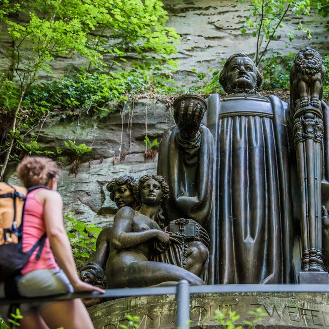 Eine Person mit Rucksack betrachtet das Richard-Wagner-Denkmal in einer Felsnische, umgeben von Pflanzen.
