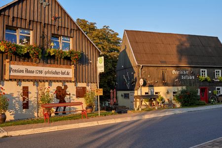 Zwei rustikale Gebäude mit Holzfassade, umgeben von Pflanzen und Sitzgelegenheiten im Freien.