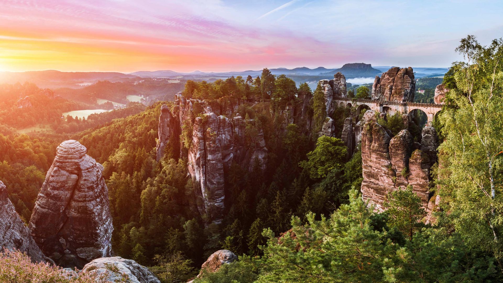 Sonnenuntergang über einer felsigen Landschaft mit Bäumen und Bergen im Hintergrund.