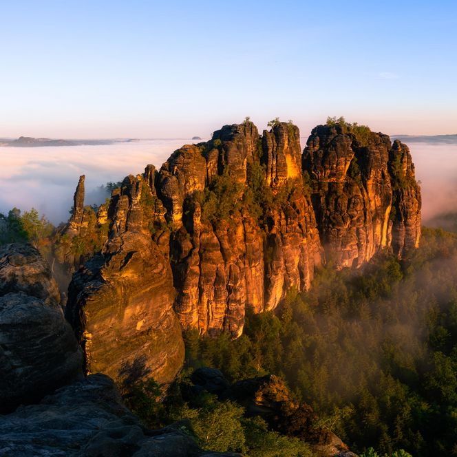 Felsformation in der Morgensonne, umgeben von Nebel und Bäumen.