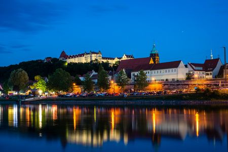Blick auf eine Stadt am Wasser bei Nacht, mit beleuchteten Gebäuden und einer Burg im Hintergrund.