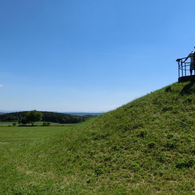 Zwei Personen stehen auf einem Hügel und blicken in die Ferne, umgeben von grünem Gras und blauem Himmel.