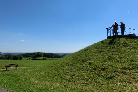 Zwei Personen stehen auf einem Hügel und blicken in die Ferne, umgeben von grünem Gras und blauem Himmel.