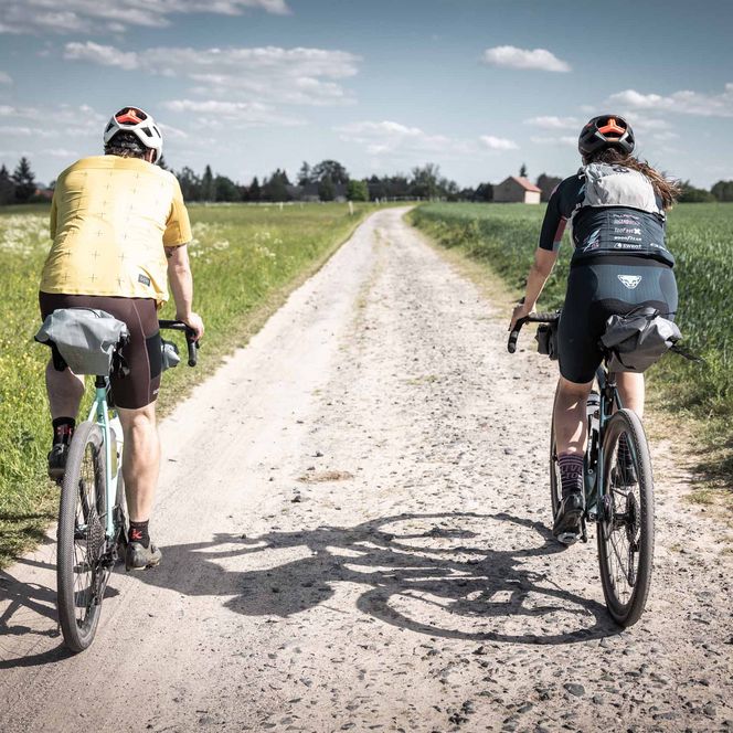 Zwei Radfahrer auf einem Schotterweg, umgeben von grünen Feldern und blauem Himmel.