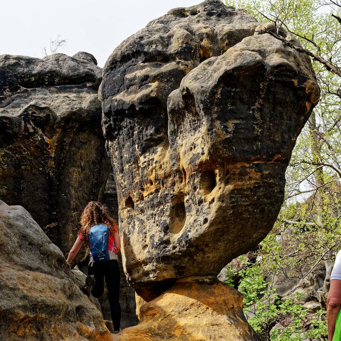 Zwei Wanderer erkunden eine Felsformation mit markanten, großen Steinen und umgebender Vegetation.