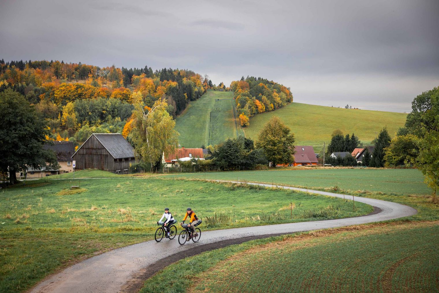 Zwei Radfahrer fahren auf einem Weg durch eine grüne Landschaft mit Bäumen und Hügeln von Rugiswalde im Hintergrund.