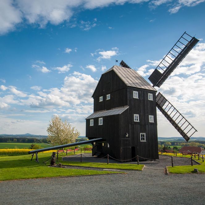 Schwarze Windmühle auf einer grünen Wiese unter blauem Himmel mit Wolken.