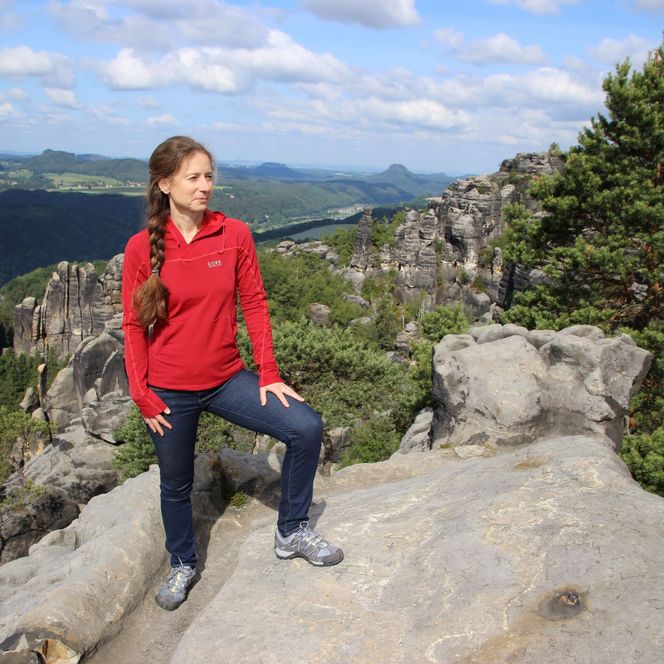 Frau in roter Jacke steht auf Felsen mit Blick auf bewaldete Hügel.