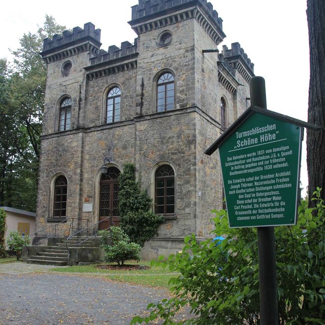 Historisches Schloss mit Türmen, umgeben von Bäumen und einem Weg. Ein grünes Schild steht im Vordergrund.
