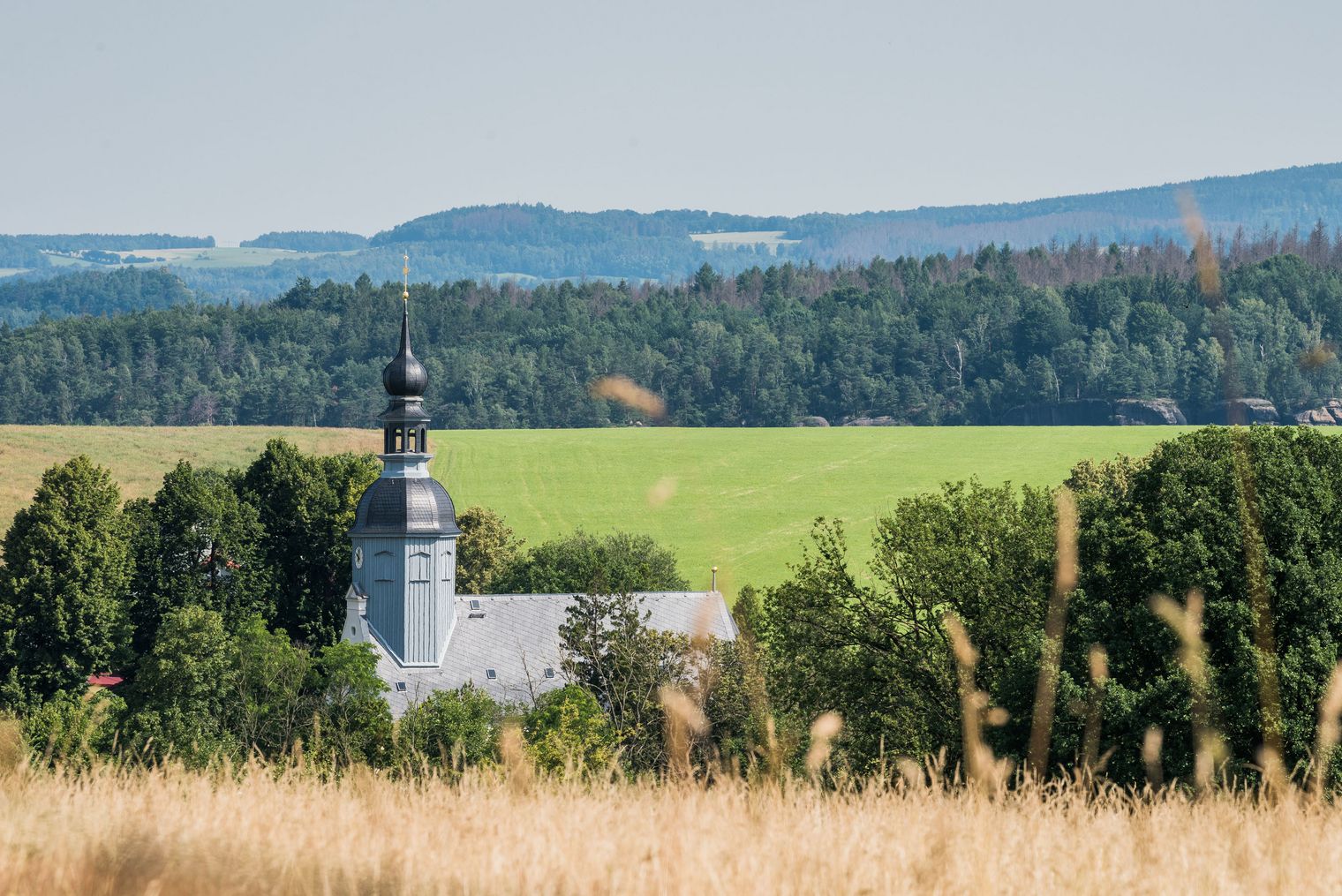 Kirche mit Turm, umgeben von Wiesen und Wäldern, im Hintergrund sanfte Hügel.