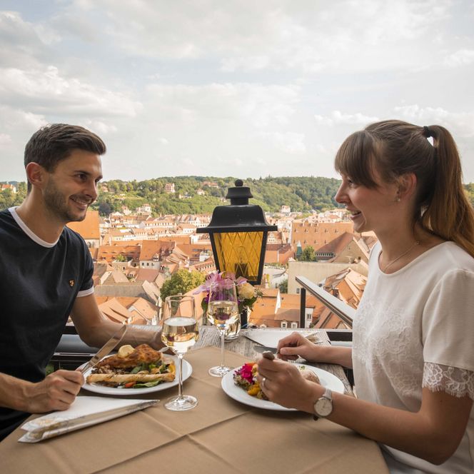 Paar sitzt an einem Tisch im Freien und genießt Essen mit Blick auf eine Stadtlandschaft.