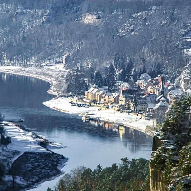Winterlandschaft mit schneebedeckten Bäumen, einem Fluss und einem kleinen, verschneiten Dorf am Ufer.