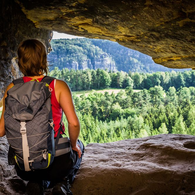 Wanderer mit Rucksack sitzt in einer Höhle und blickt auf eine grüne Landschaft mit Bäumen und Bergen.