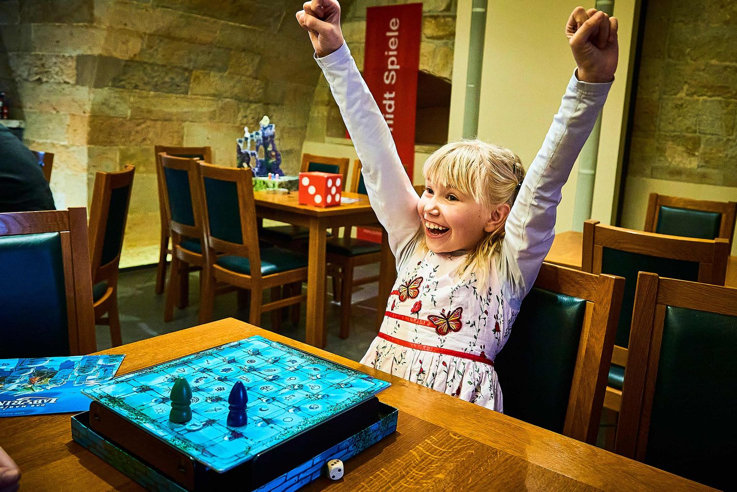 A girl with blonde hair raises her arms in delight in front of a chessboard.
