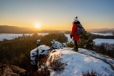 Person in roter Jacke steht auf einem schneebedeckten Felsen und schaut auf den Sonnenuntergang über einer winterlich...